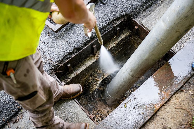 A plumber's water jet blasting silt out of a drain chamber, with a suction hose beside it